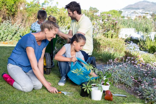 A family of four engaging in gardening activities in a well-maintained backyard garden with lush grass, a variety of flowering plants, and a neat flower bed border. The mother, dressed in a blue top and grey trousers, is kneeling on the grass, planting or tending to a small shrub. A young girl, wearing a blue top and jeans, is sitting on the grass, holding a small garden tool and a blue watering can with a plant design. The father, in a light-coloured t-shirt, is standing behind them, assisting or observing, while a young boy, dressed in a striped shirt, is nearby. The garden features a paved pathway or patio area partially visible, with garden tools and watering cans arranged nearby. The background shows more flowering beds, shrubbery, and trees, with a scenic view of distant hills and a partly cloudy sky, indicating a bright, outdoor environment. The scene suggests regular outdoor maintenance, gardening, and family activity aligned with professional garden care services in Fulham, contributing to a vibrant and well-kept outdoor space.