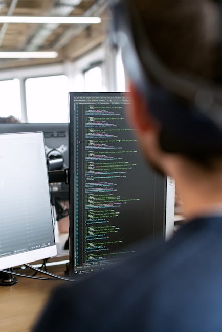 The image shows the back of a person working at a desk in an industrial-style office with large windows allowing natural light. The individual is focused on a computer monitor displaying lines of code